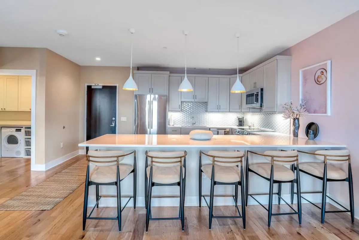 A modern kitchen scene with a central island, comfortable barstools, decorative items on the counter, and visible appliances. The layout encourages casual dining and mingling, with lighting and décor that enhance the cozy, contemporary feel.