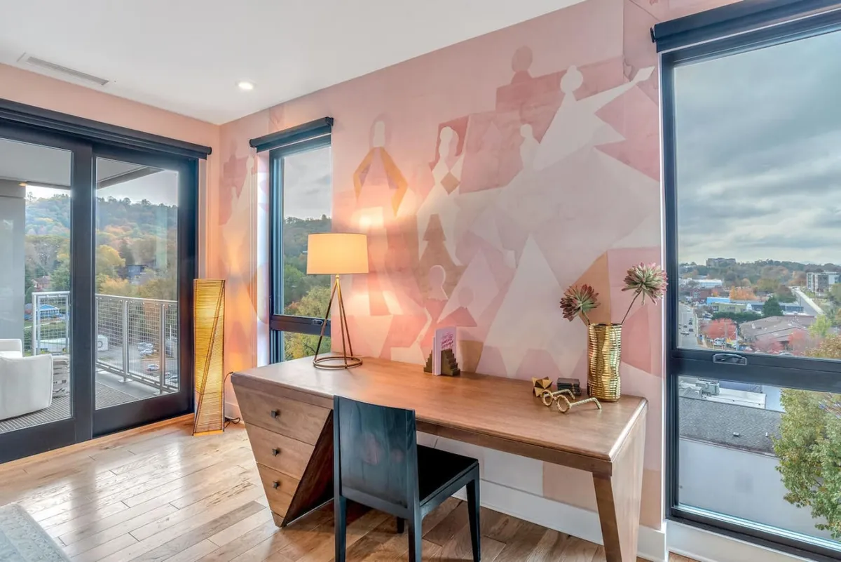 A stylish workspace set against large windows that let in plenty of natural light. The area features a wooden desk, artistic wall mural, and table lamp, with sweeping outdoor views visible through the glass. This scene emphasizes the deep connection between interior comfort and the surrounding landscape.