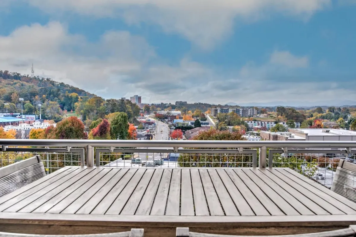 This view image captures a wooden outdoor table set against a cityscape with autumn foliage and distant mountains. The vantage point offers a glimpse of the city’s vibrancy and the scenic surroundings, blending urban excitement with natural beauty.