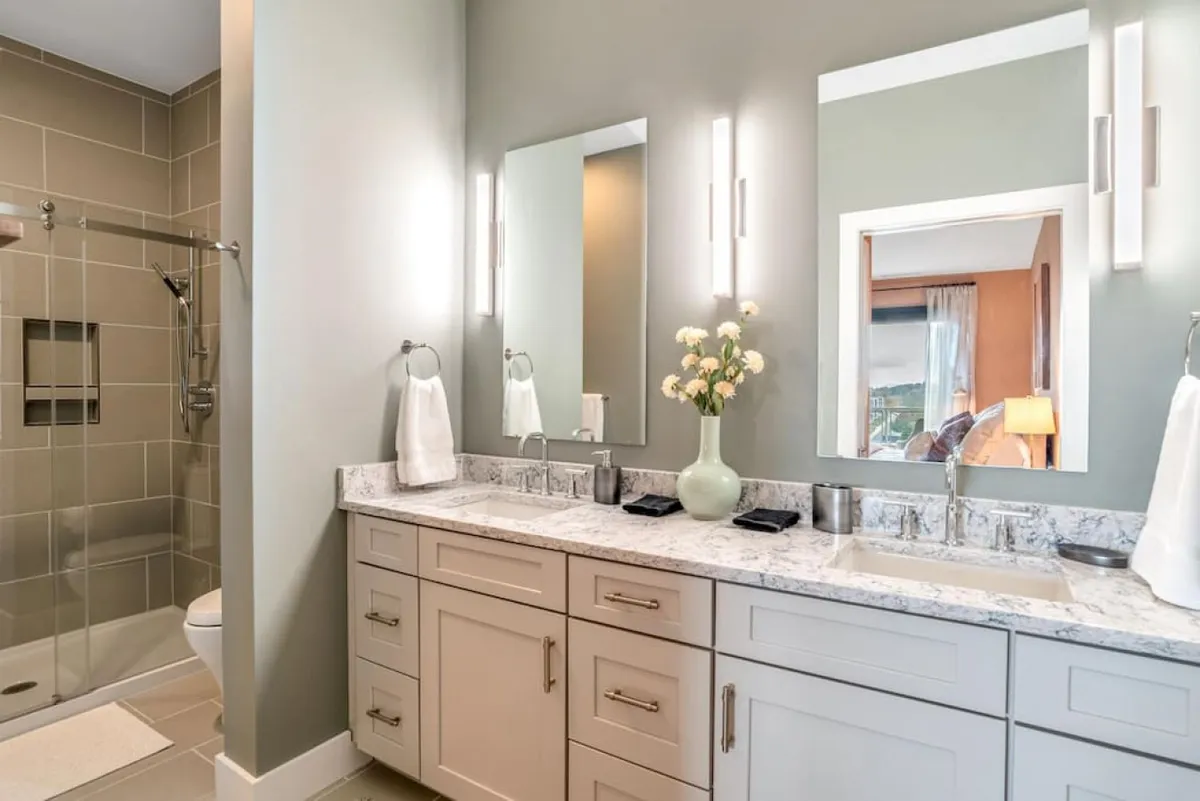 A serene bathroom with a spacious vanity, dual mirrors, a shower area, and a vase with fresh flowers. The elegant design and thoughtful details evoke a peaceful and rejuvenating environment perfect for morning and evening routines.