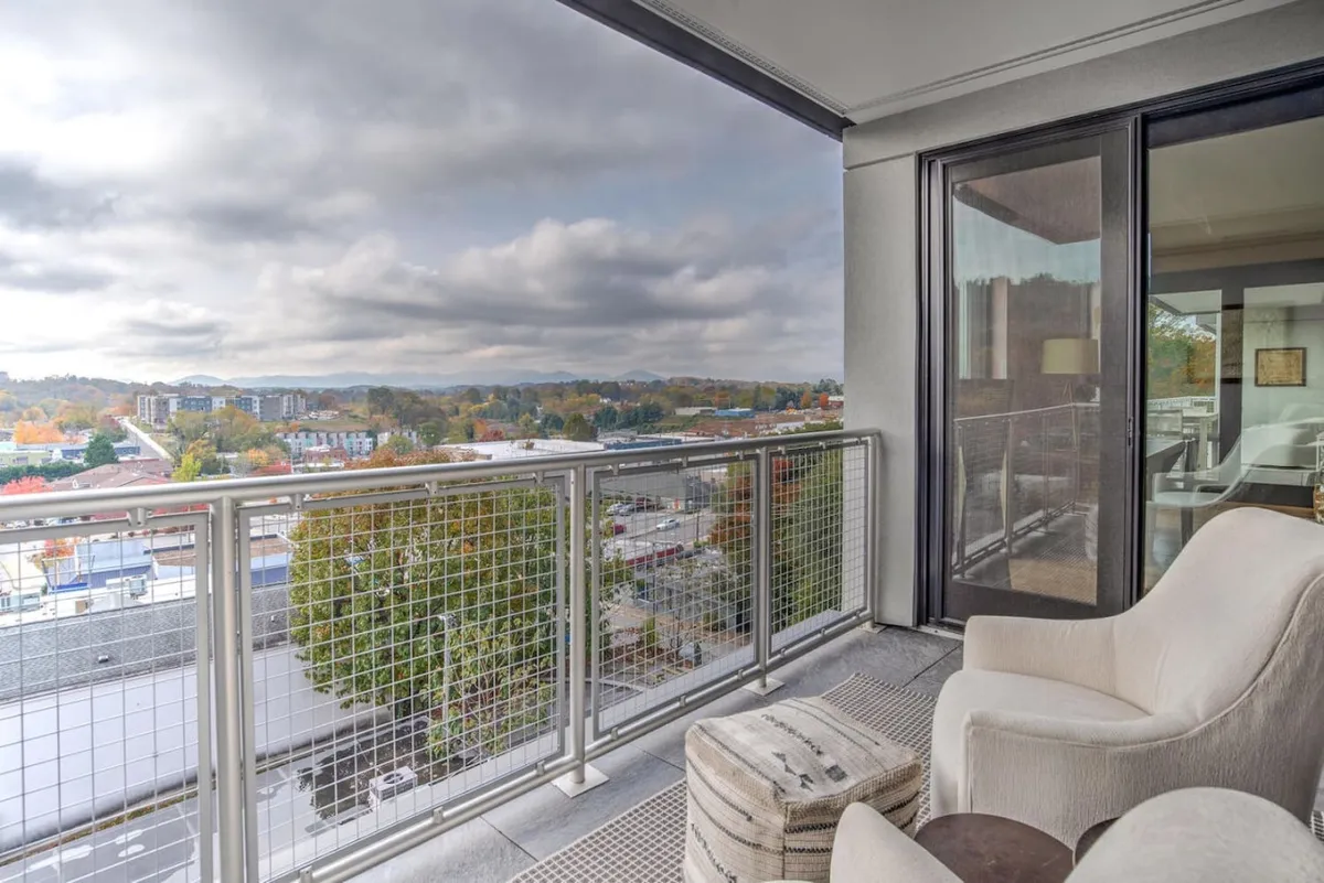 This exterior image captures a comfortable balcony with a textured ottoman and a chair, set against an expansive view of the landscape beyond downtown Asheville. The inviting setup suggests a perfect transition space between the condo and the city, with a serene atmosphere for morning coffee or evening relaxation.