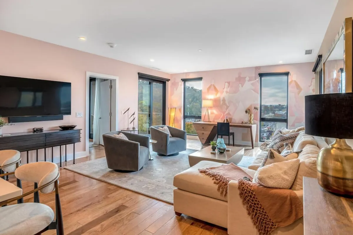 A bright living area featuring a plush sofa, comfortable armchairs, a modern coffee table, and tasteful wall art. Large windows in the background offer a clear, unobstructed view of the mountains, capturing the interplay of natural light and inviting warmth into the room. The space blends comfort and style, perfectly illustrating an open-concept living area designed for both relaxation and social gatherings.