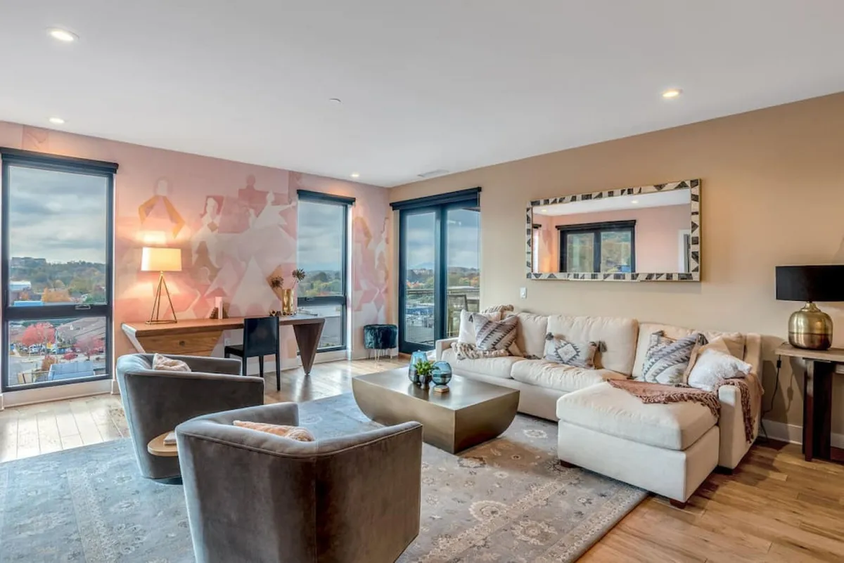 The hero image showcases a stylish living room with a modern sofa, a decorative mirror reflecting natural light, an artistic wall mural adding a creative flair, a sleek coffee table, and inviting hardwood flooring. The space is bathed in soft morning light, hinting at the sunrise views visible from the windows.
