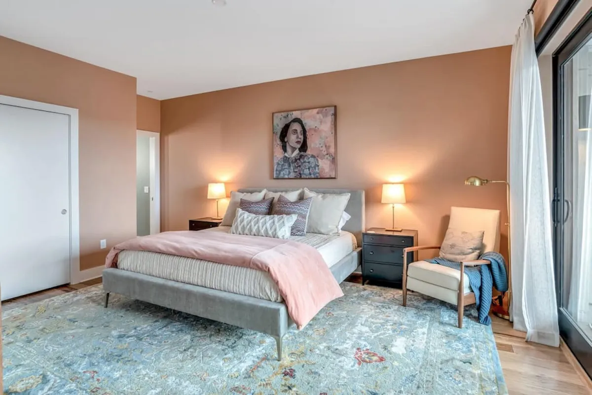 A tranquil bedroom setting decorated with soft, decorative pillows, warm lighting from nightstand lamps, thoughtful artwork, and an accent chair near the window. The arrangement suggests enjoying peaceful mornings with natural light and possibly glimpses of the scenic outdoors.