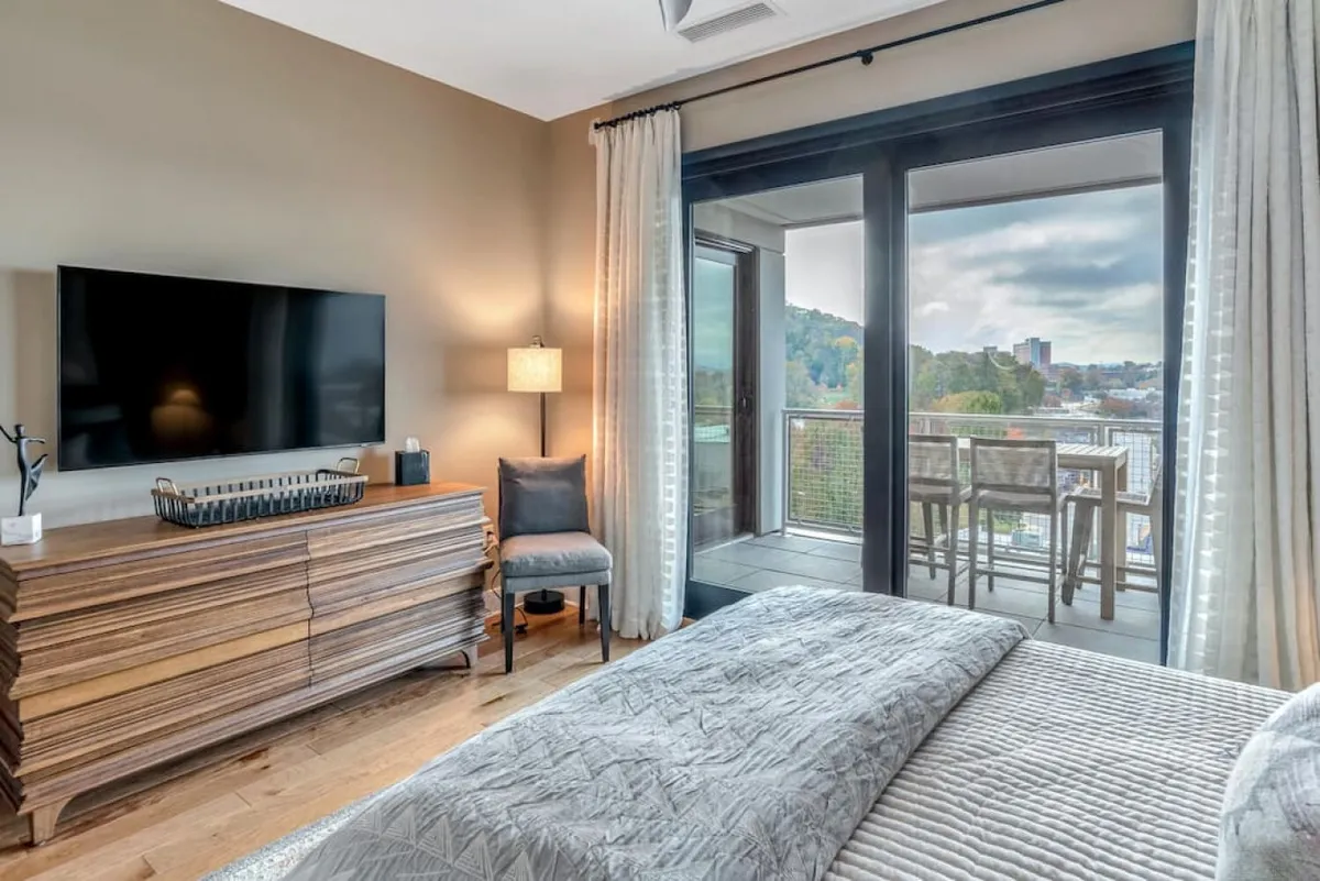 This tranquil bedroom features a plush bed with textured bedding, a wooden dresser, and a lamp on the bedside table. The large window fills the room with natural light and offers refreshing views, reinforcing the room’s relaxing atmosphere.