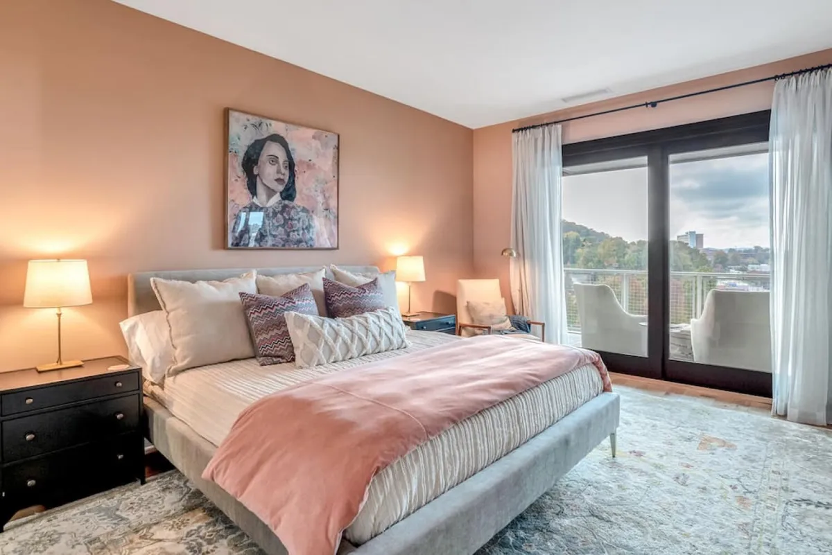 A cozy bedroom featuring a comfortable bed positioned to take advantage of the window's view, with carefully placed artwork above the bed and soft natural light filling the space. The window frames a scenic view—potentially mountains or cityscape—implying a strong connection to the outdoors.