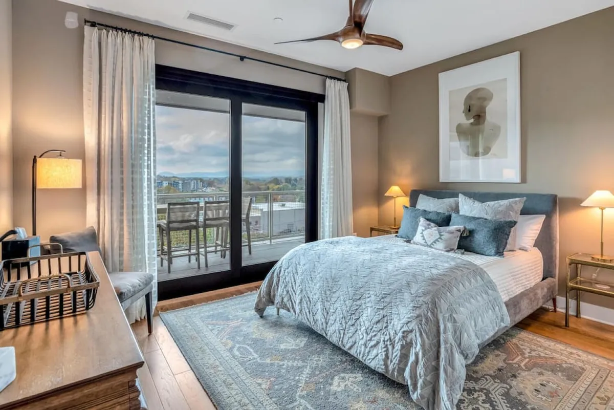 A serene bedroom with a blue quilt and decorative pillows on the bed, complemented by large glass doors leading out to a balcony. Artwork and wooden furniture add a sophisticated touch, while the abundant natural light emphasizes spaciousness and tranquility.