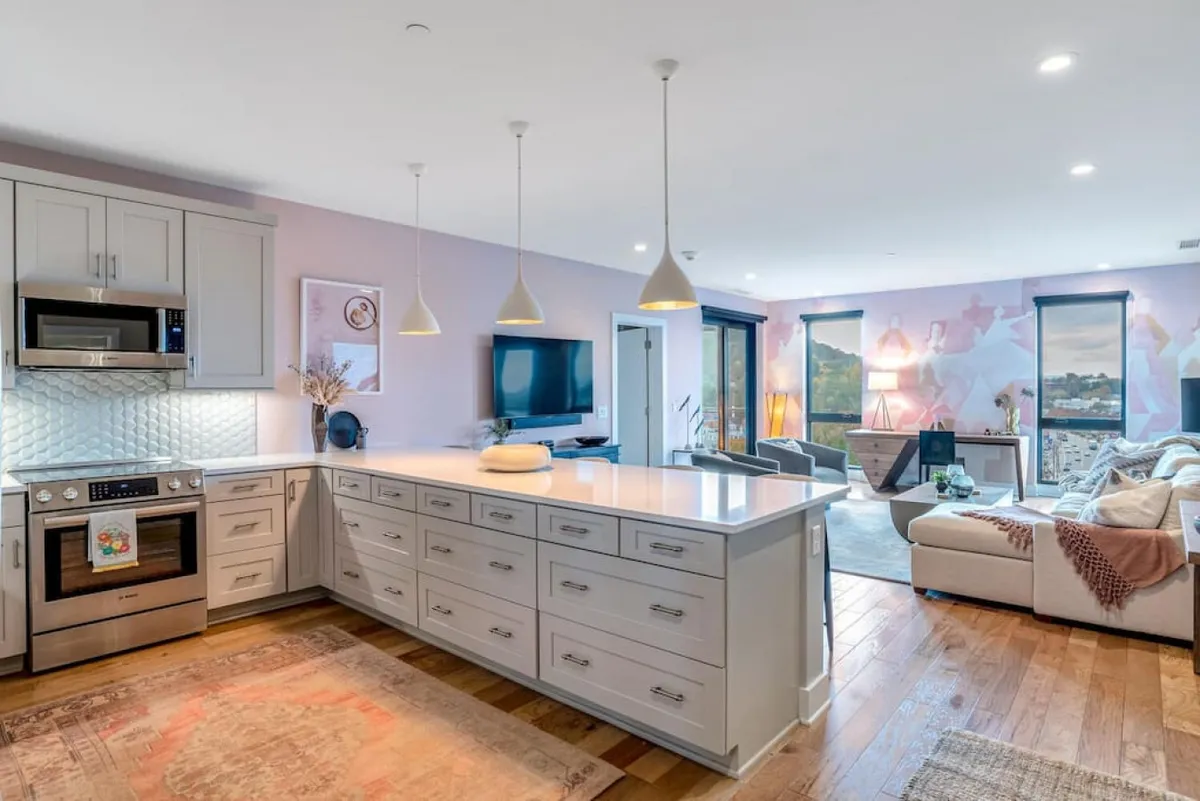 A welcoming open-plan living area featuring a comfortable sofa, a kitchen with modern appliances, and an inviting island countertop. Bright artwork on the wall adds a playful touch, and the space is well-lit and designed for communal activities like cooking, gathering, and relaxing together.