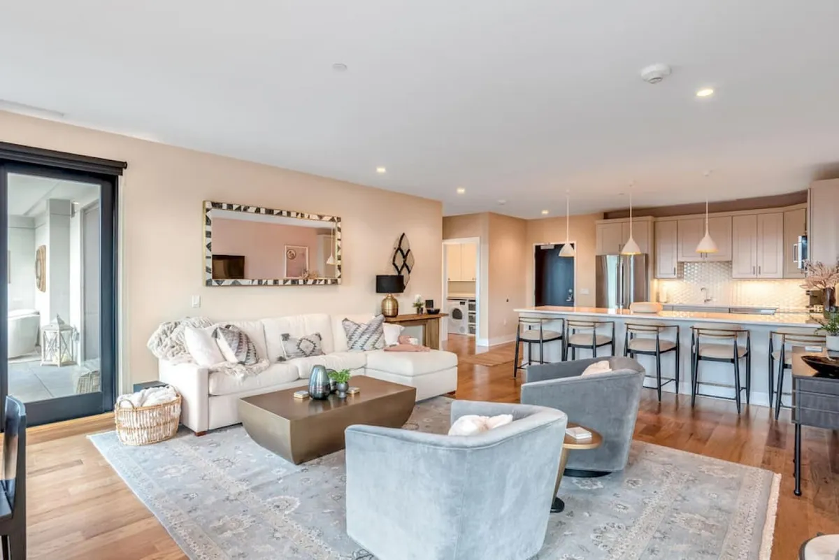 A spacious, sunlit interior featuring a contemporary sofa, sleek coffee table, inviting kitchen island with bar stools, and mirrored accents. The layout highlights the open-concept design, seamlessly connecting living and kitchen spaces. Natural light fills the room, reinforcing the sense of space and connection to the outdoors.
